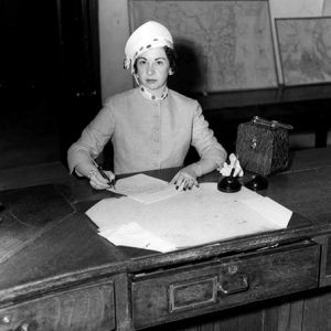 White woman signing paper at desk in room with maps leaning against the wall behind her