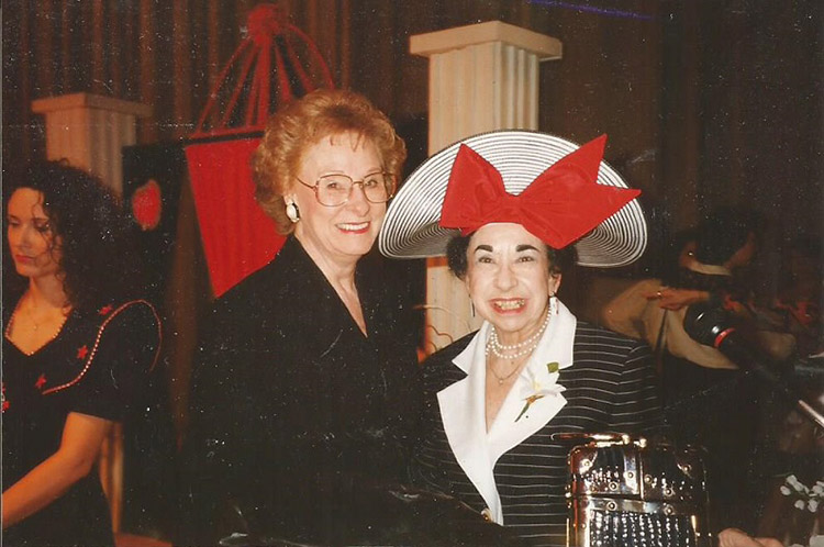 White woman with glasses in dress standing next to white woman with large hat with bow tie on it