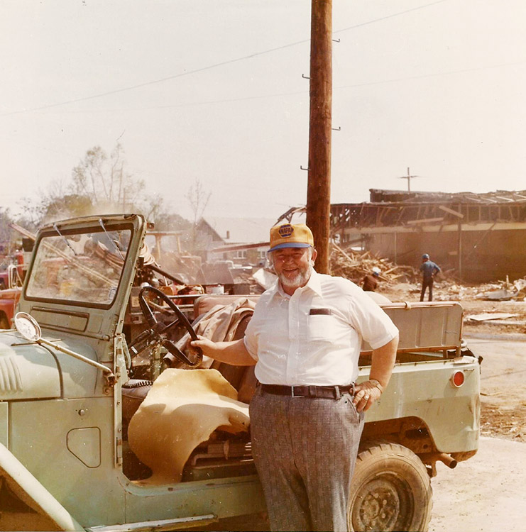 Older white man with hat standing next to Jeep at site of damaged house