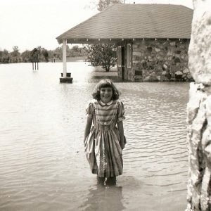 Young white girl smiling on flooded street with single-story stone building with covered porch behind her