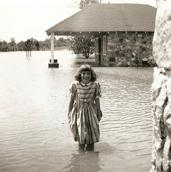 Young white girl smiling on flooded street with single-story stone building with covered porch behind her