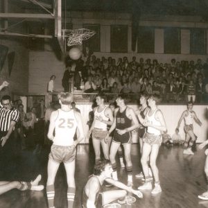 White referee making a call during a basketball game in school gymnasium surrounded by young white players
