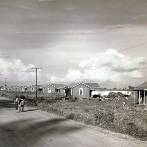 Dirt road with row of small identical houses and power lines on the right side with two children standing in road