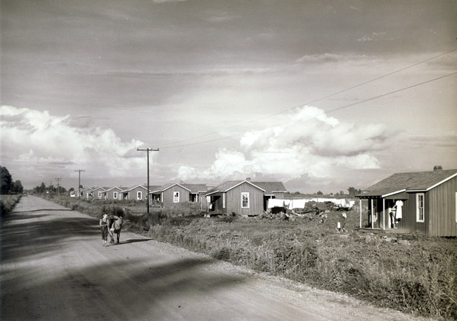 Dirt road with row of small identical houses and power lines on the right side with two children standing in road