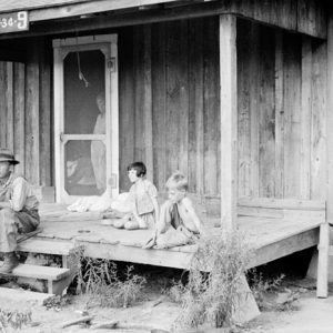 White man and two children sitting on porch of wooden cabin with woman looking through out from the screen door