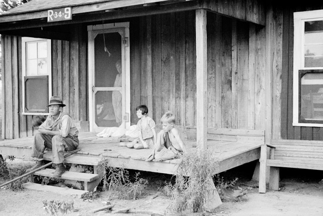 White man and two children sitting on porch of wooden cabin with woman looking through out from the screen door