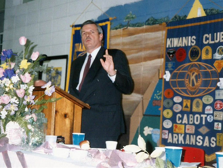 White man in suit speaking at lectern with flowers in the foreground