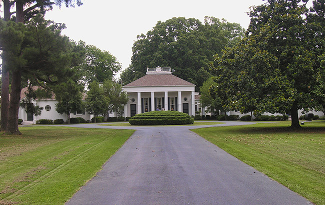 Mansion house with covered porch and single-story wings on estate with paved driveway and trees