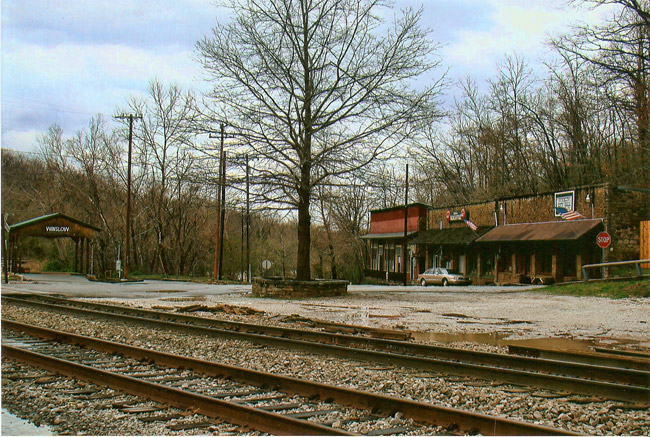 Three brick storefronts with covered porches and covered bridge on street with pair of railroad tracks in the foreground