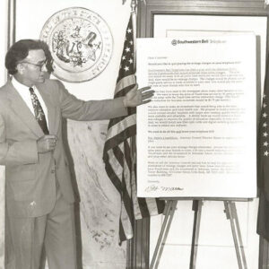 White man with glasses in suit and tie explaining poster size document on easel between flags