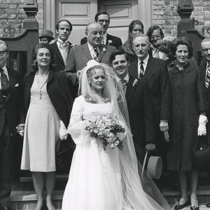 Young white woman in wedding dress holding flowers and young white man in suit with white men and women standing behind them