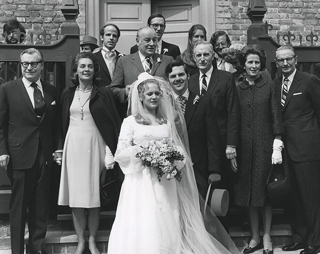 Young white woman in wedding dress holding flowers and young white man in suit with white men and women standing behind them