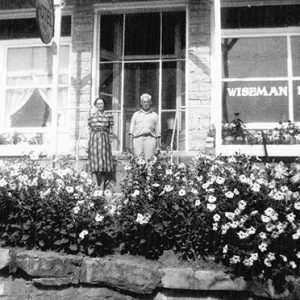 Old white man and woman standing outside hotel with flowers and brick wall