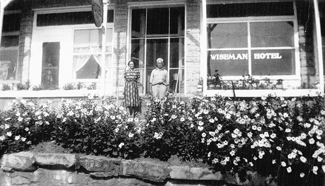 Old white man and woman standing outside hotel with flowers and brick wall