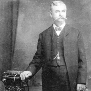 Older white man with long mustache in three-piece suit and bow tie with his right hand on a typewriter