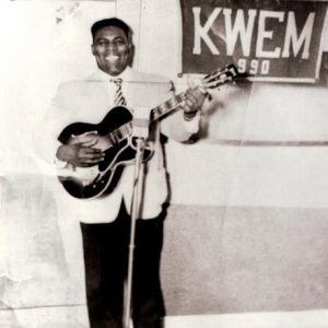 African-American man smiling with guitar and "K.W.E.M. 990" banner hanging on the wall behind him