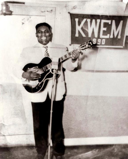 African-American man smiling with guitar and "K.W.E.M. 990" banner hanging on the wall behind him