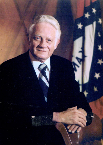 Old white man smiling in suit and tie with Arkansas flag behind him