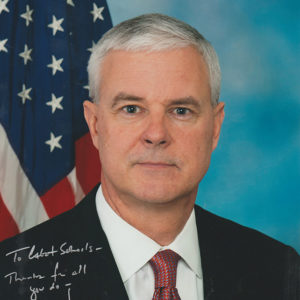 White man wearing suit and tie in front of flag
