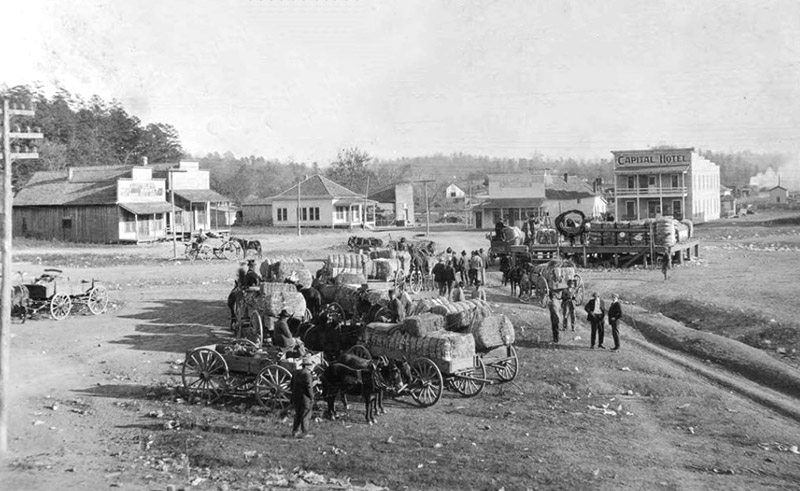 Men with horse drawn wagons loaded with cotton in town with dirt roads and buildings in the background