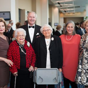 Older white man in tuxedo standing and smiling with group of white women in hallway