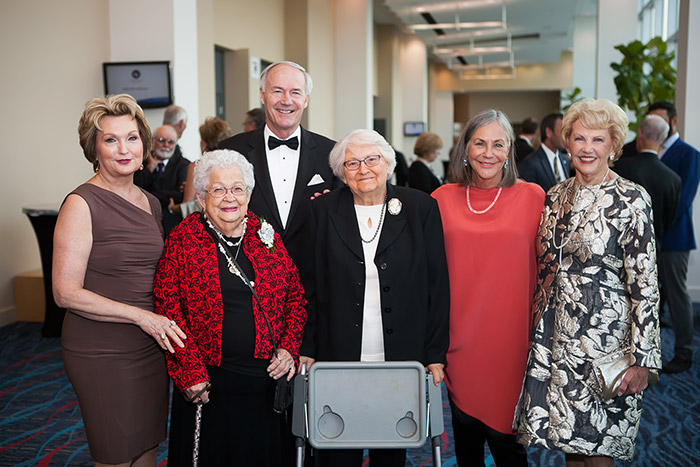 Older white man in tuxedo standing and smiling with group of white women in hallway
