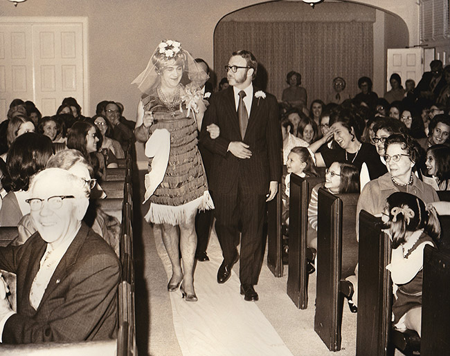 White man in dress and veil with man in suit in crowded church sanctuary