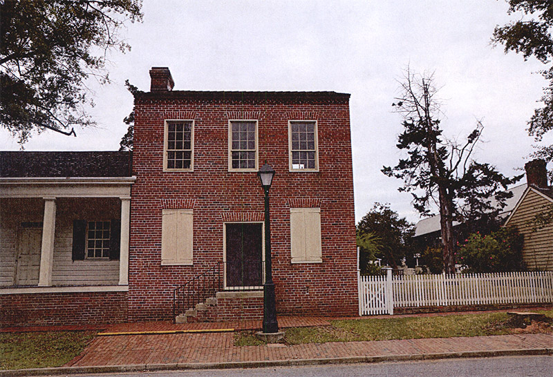 Front view of single-story wooden structure with covered porch adjoining two-story brick building on street
