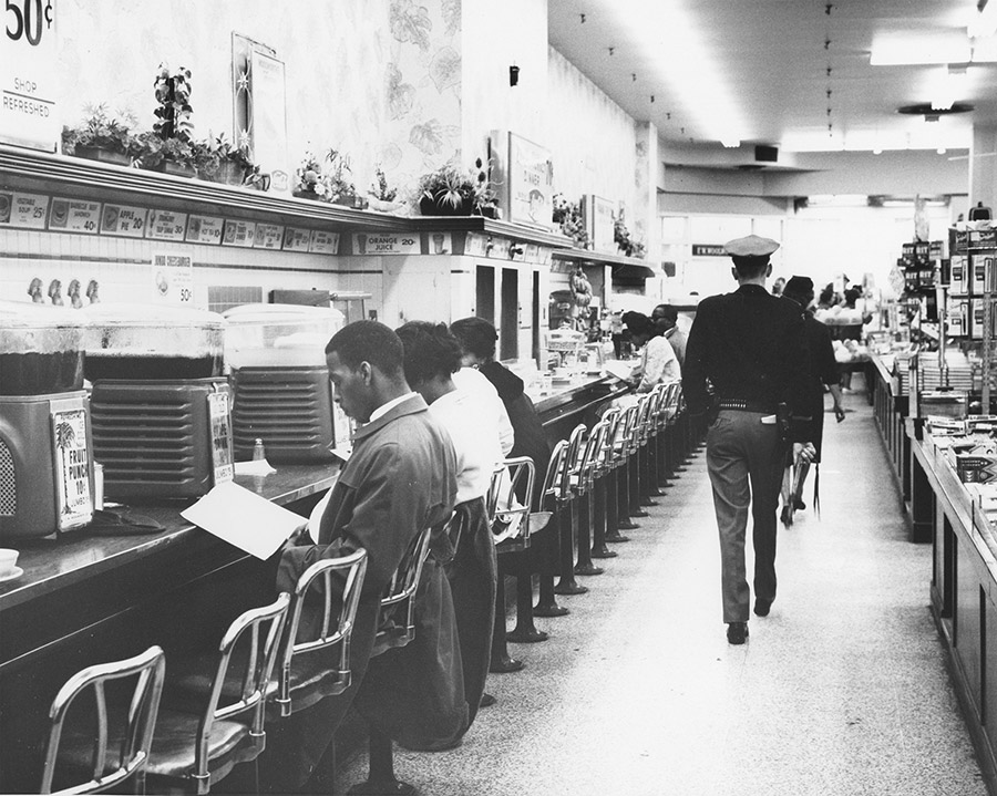 Black students sitting at lunch counter with white policeman walking by them