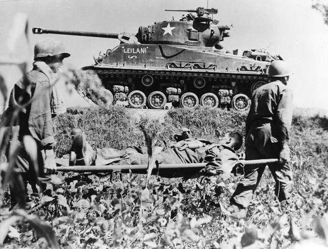 White soldiers carrying white man on stretcher with tank behind them