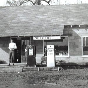 White man standing outside single-story building with Post Office sign and two gas pumps on dirt parking lot