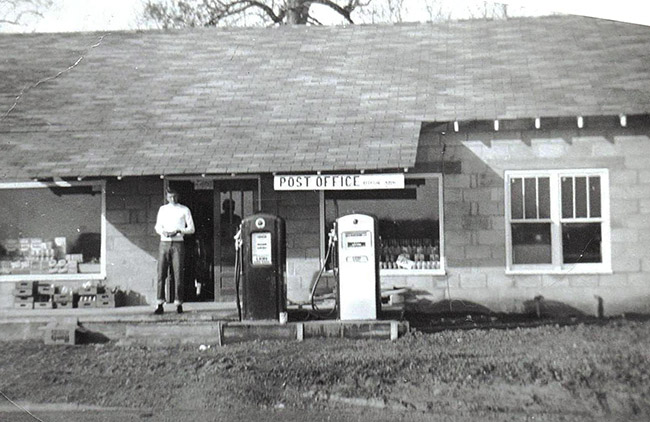 White man standing outside single-story building with Post Office sign and two gas pumps on dirt parking lot