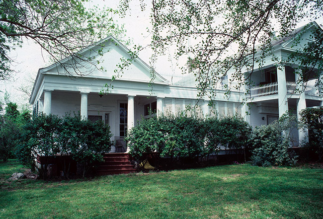Two-story house with covered columned porch and balcony and single-story wing with its own entrance and porch with bushes in front