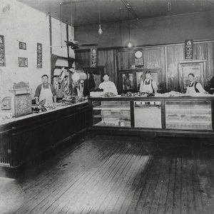 White men in aprons standing behind counter displaying cut meat