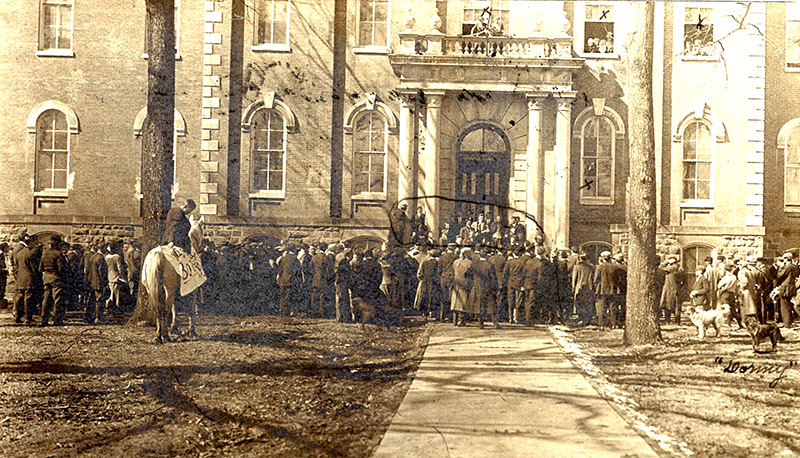 Crowd of white men women and white man on horseback outside multistory building with balcony and arched windows