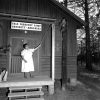 Woman ringing bell from wood frame porch with sign "Yale Forestry Camp Crossett - Arkansas"