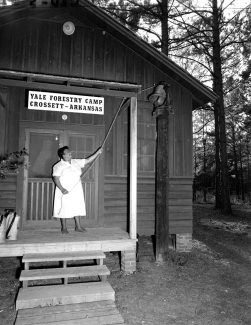 Woman ringing bell from wood frame porch with sign "Yale Forestry Camp Crossett - Arkansas"