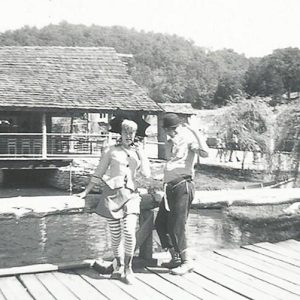 White man and woman in costume with pool and cabin behind them