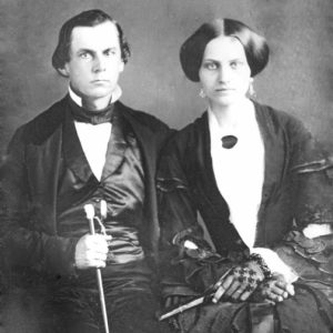 White man in suit and vest holding a cane with white woman in dress and gloves