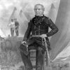 White man sitting in military uniform with sword at camp with tents behind him