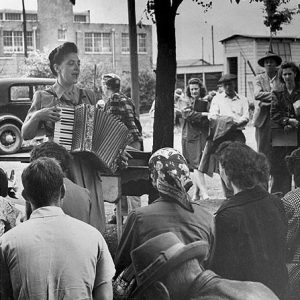 White woman singing and playing an accordion to a white crowd with car and buildings behind her