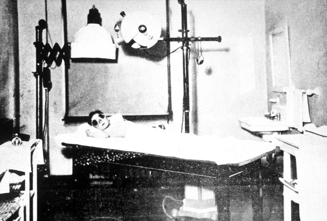 Black and white photo of white boy in tinted goggles lying on examination table below lights and medical device