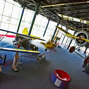 large interior space with floor to ceiling windows and various old aircraft and automobiles hanging from ceiling and positioned on the floor