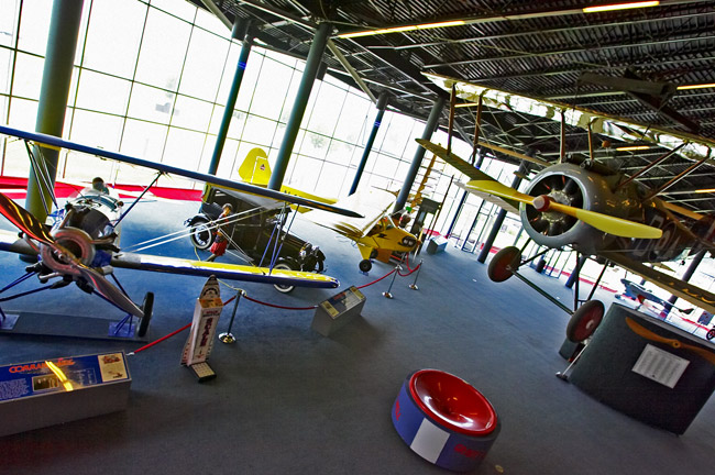 large interior space with floor to ceiling windows and various old aircraft and automobiles hanging from ceiling and positioned on the floor
