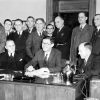 Group of white men in suits standing behind three suited white men sitting at a desk