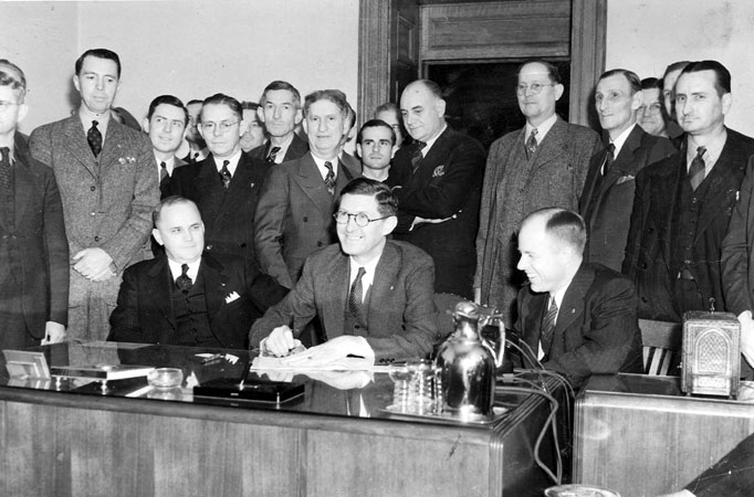Group of white men in suits standing behind three suited white men sitting at a desk