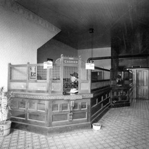 White men in hat and suit sitting behind a long cashier's desk with bars and stove pipe