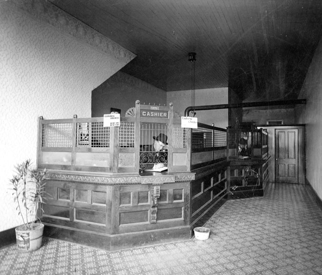 White men in hat and suit sitting behind a long cashier's desk with bars and stove pipe