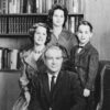 White man in suit sitting in chair before wife, son, and daughter, with bookshelves behind them