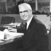 White man with glasses in suit and tie sitting at his desk